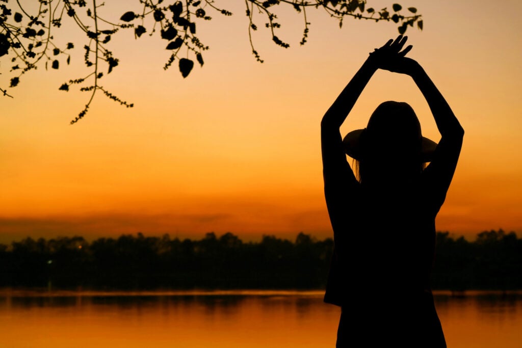 Silhouette of a woman with her hands in the air by a lake. Different shades of orange surrounds her.