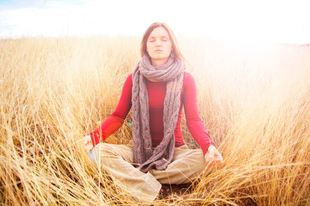 Woman meditating in an open field in the long autumn grass with a white aura surrounding her.