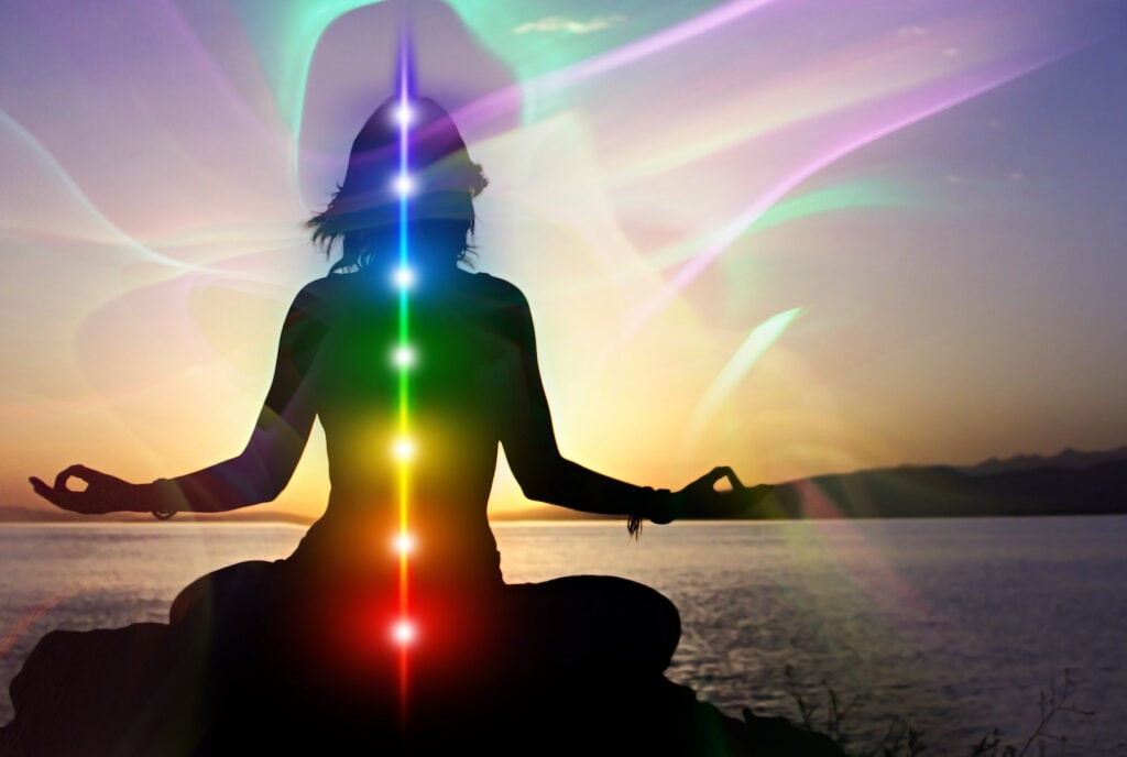 Silhouette of woman in lotus pose on the beach with the seven chakras showing while she is surrounded by aura colors.