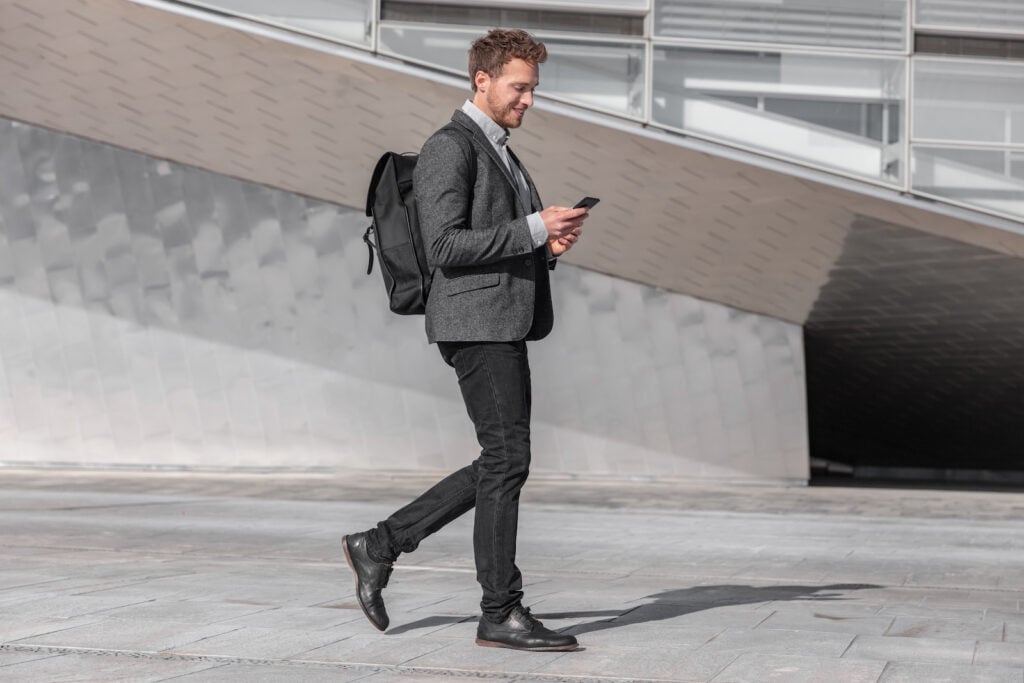 Happy young business man using phone walking with bag on city street. 