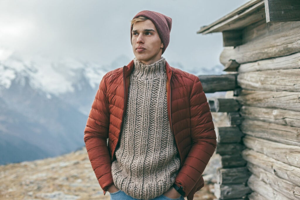 Man wearing warm sweater, winter coat, and hat posing behind mountains with snow.