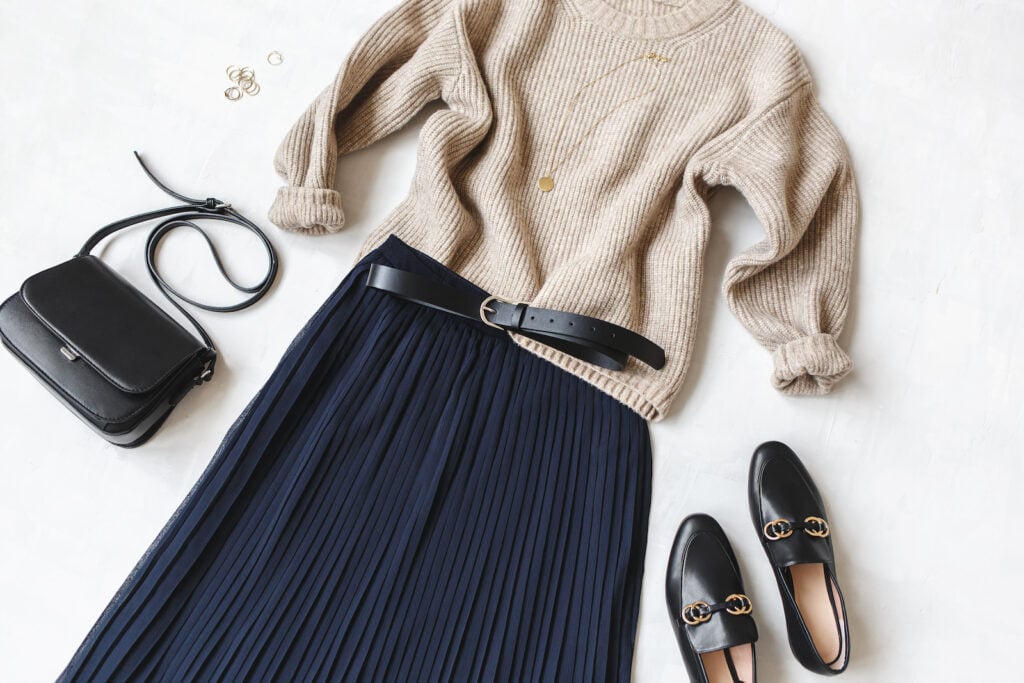 Blue midi pleated skirt, beige knitted sweater, and small black cross body bag.