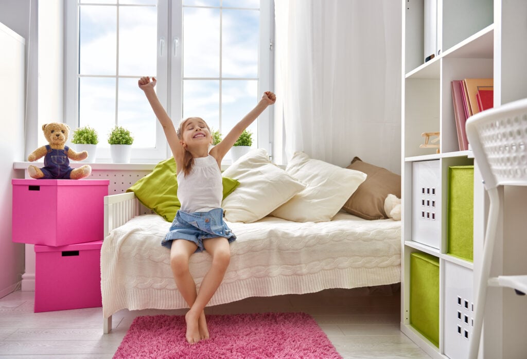 Girl sitting on bed in room with pink rug and boxes.