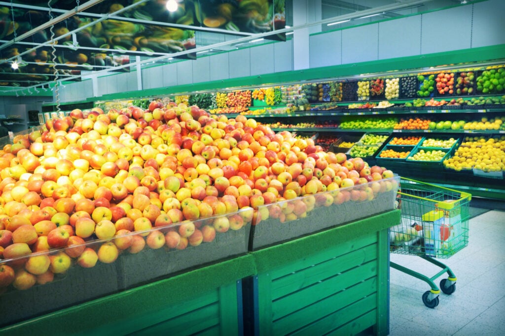 Interior of grocery store with fresh fruits and vegetables, green displays, and green shopping cart