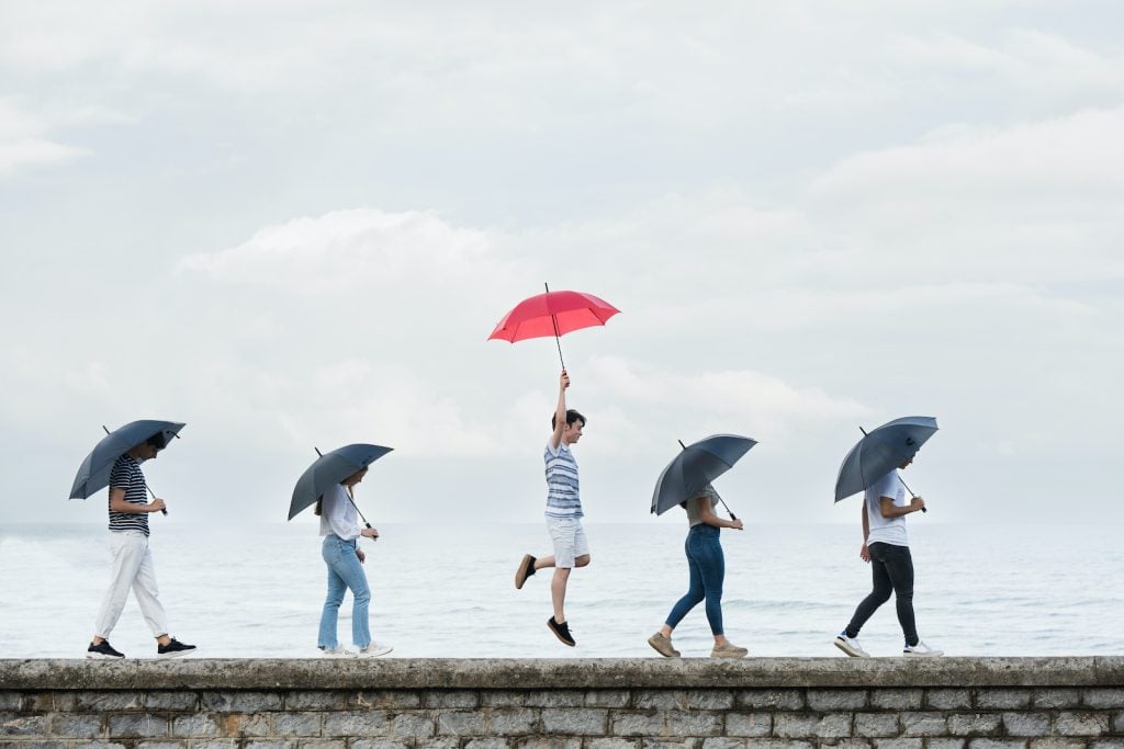 A jubilant boy holds a red umbrella, standing out from a sad line of black umbrellas