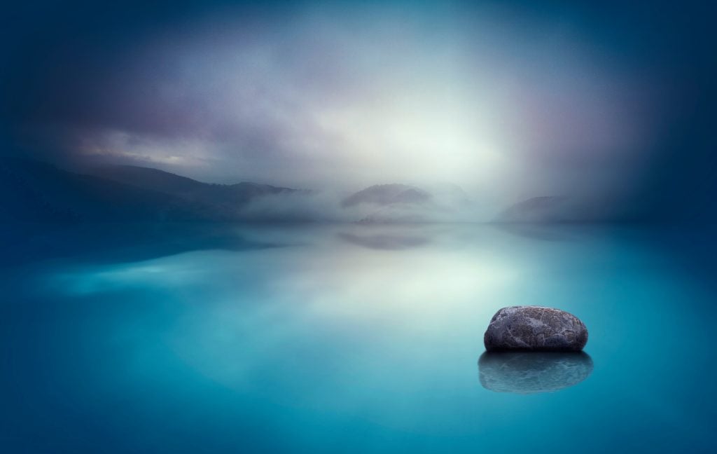 A dreamy landscape with still blue water and a rock in the foreground