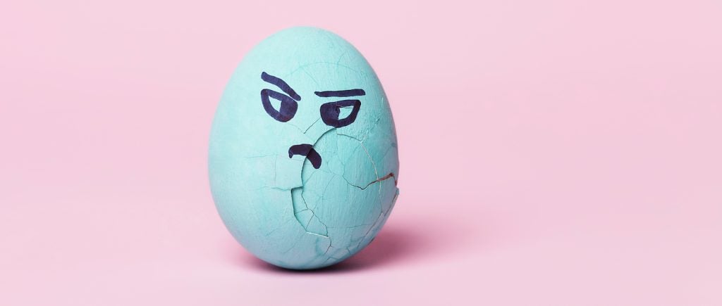 A cracked turquoise egg with a grumpy face stands on a pastel pink background