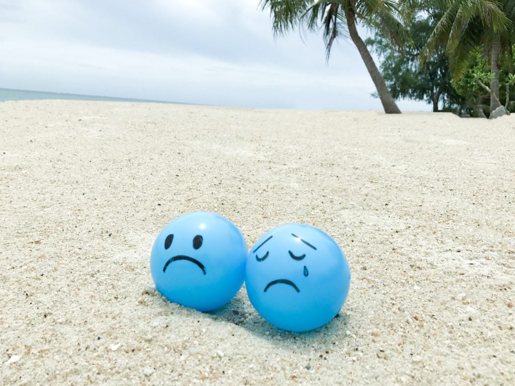 Two sad-faced blue beach balls rest on the sand at a tropical beach