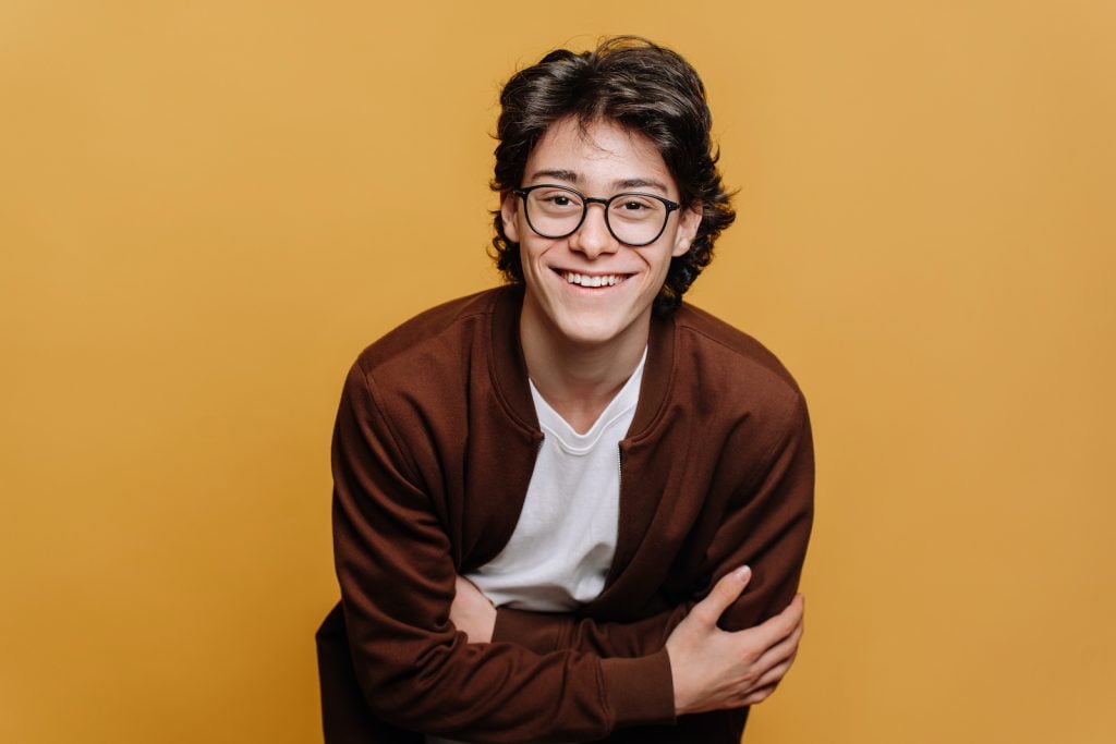 A young, smiling man wearing a brown sweatshirt and glasses against a yellowish background