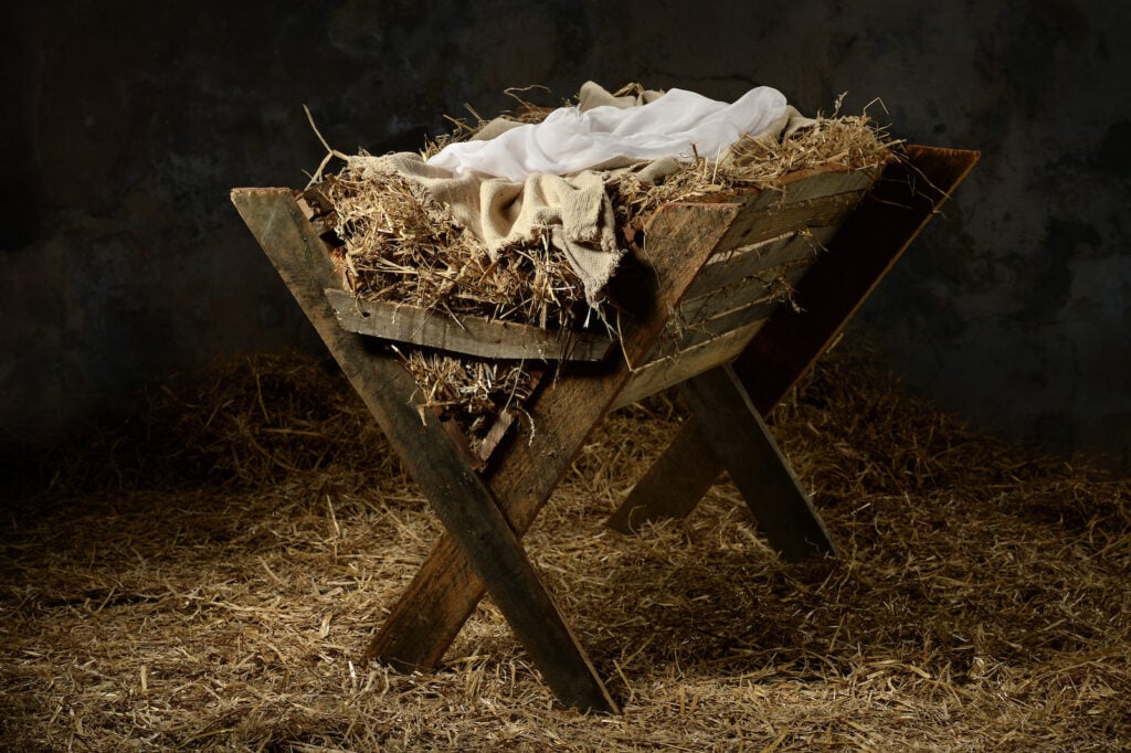 A brown wooden manger with blankets and straw stands in a stable