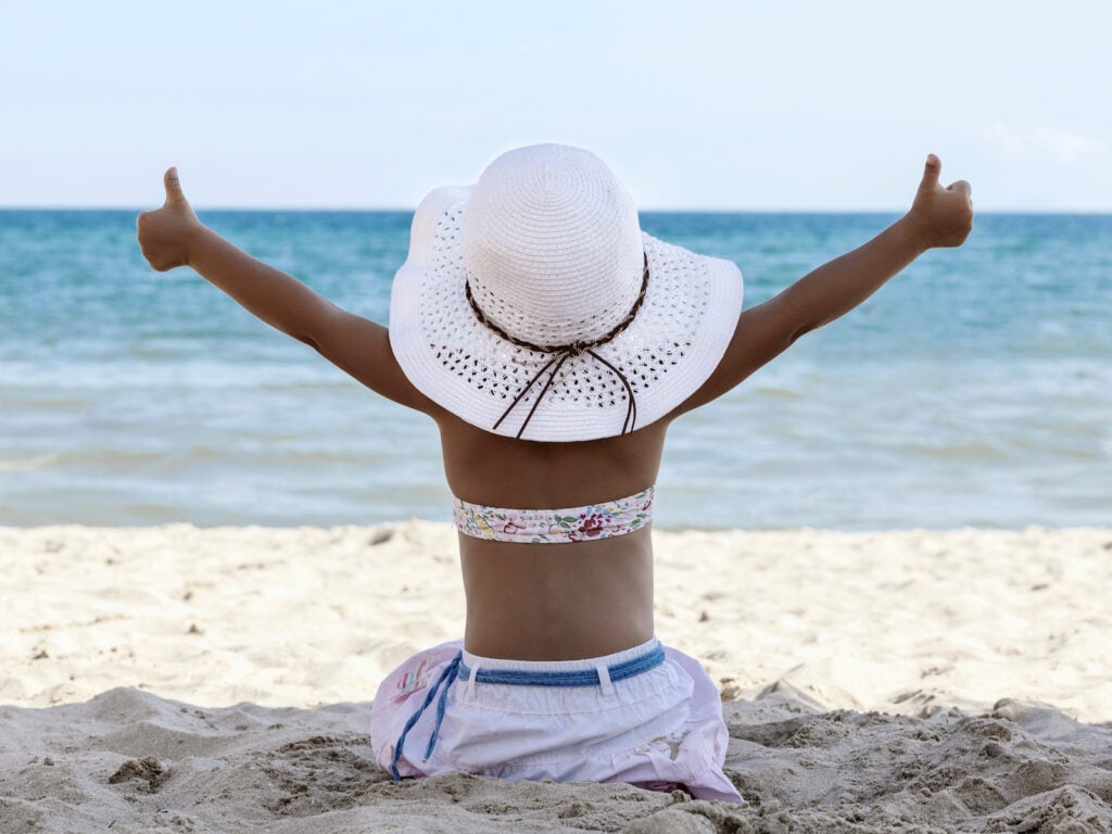 A happy child wearing a white sun hat sits facing the ocean