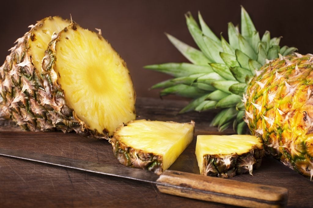 One whole ripe pineappel and one cut in slices lies on a rustik dark brown table