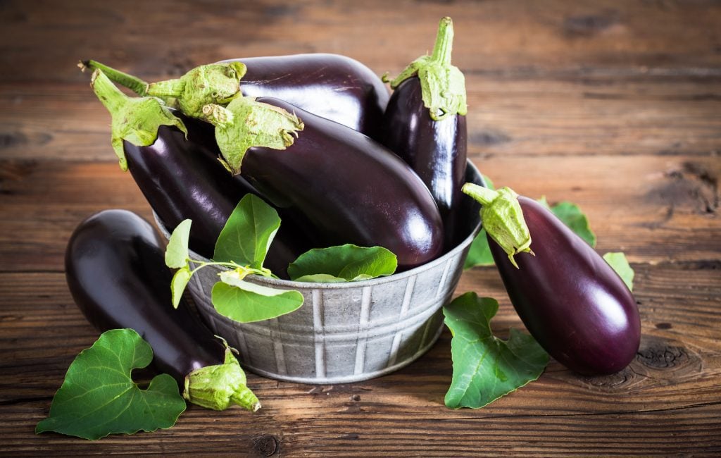 Bunch of fresh eggplants in a metal basket standing on a dark wooden table