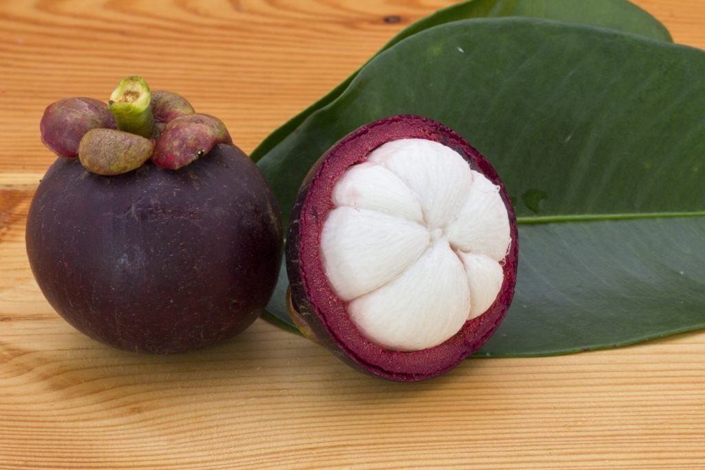 Ripe mangosteens one half mangosteen and one whole lying next to two green leaves on a wooden table