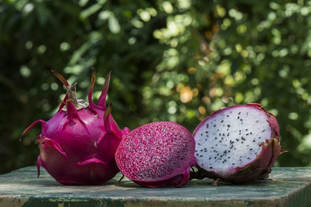 Two half of a purple and white flesh dragonfruit lies beside a whole dragonfruit on an rustic wooden table with green blurred background