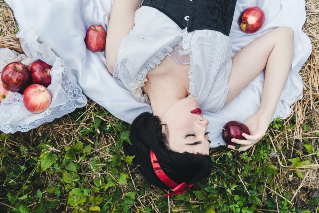 Snow White from the fairy tale is laying on the ground deep asleep surrounded by red apples