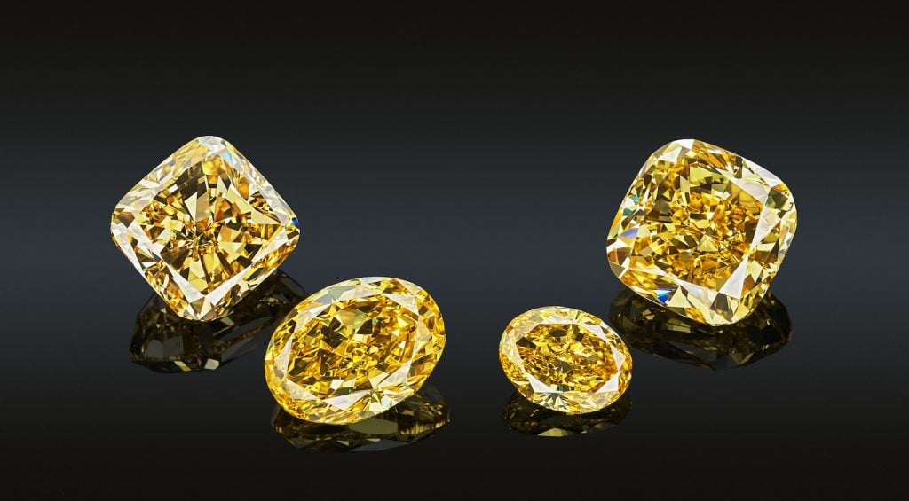 Closeup of 4 yellow diamonds in two different shapes, sizes and cuts reflected on black glass they are placed on 